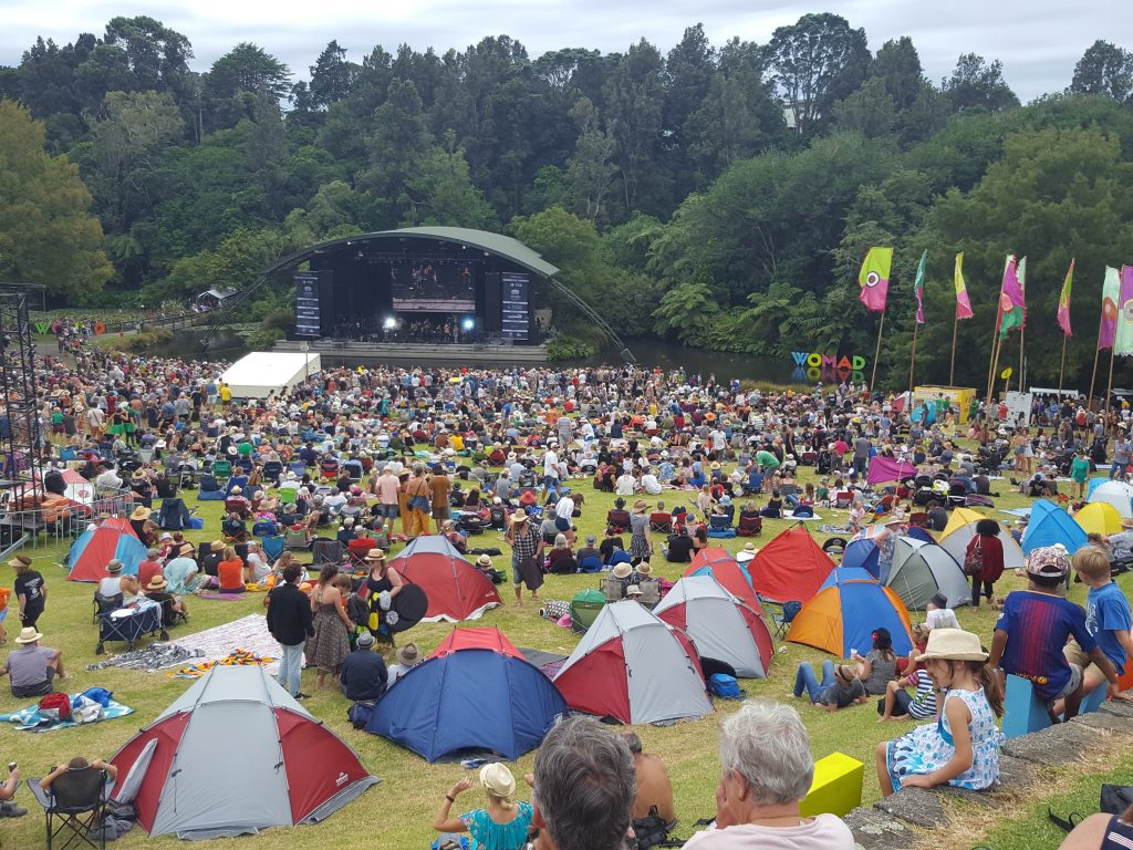 an open air concert with a stage in the distance and trees behind it. People are sitting on the grass in groups some under sunshelters