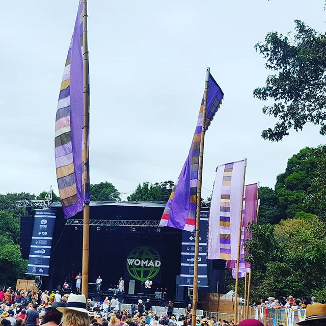 a stage in the open air set in parkland. You can see the trees in the background, the sky is pale blue, tall flags of different colours fly in the breeze, the stage is black and there are people standing and sitting a in front of it.
